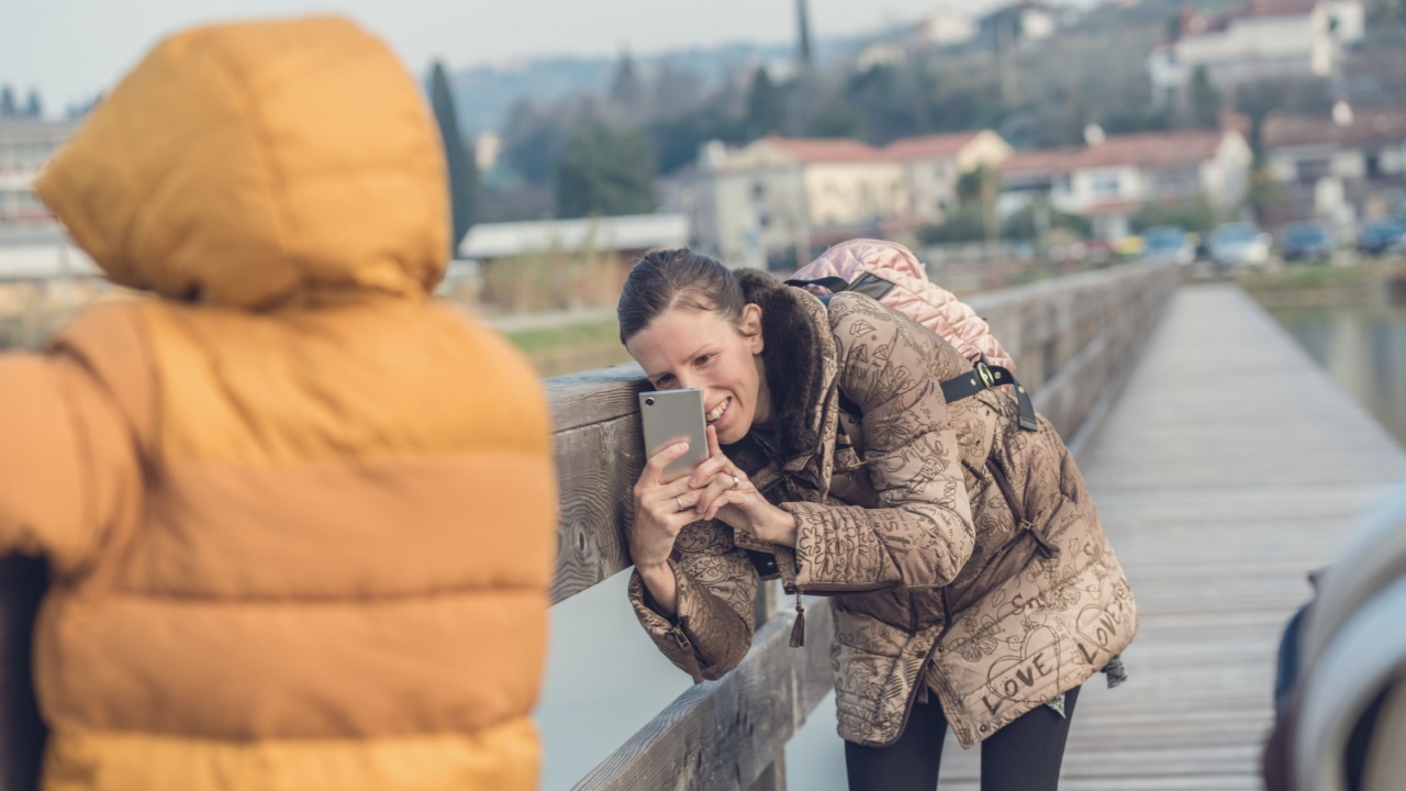 Happy young mother taking a photo with her mobile phone of her toddler child outside on a wooden path by the sea.