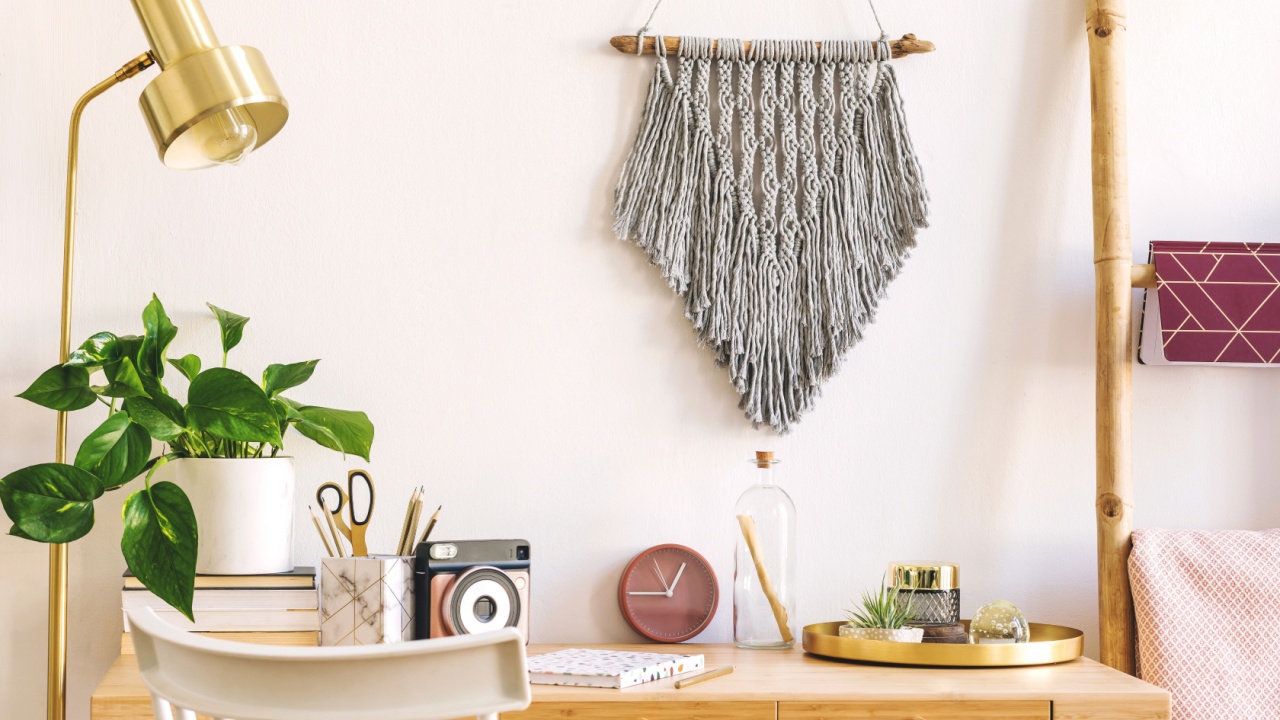 Stylish scanidnavian interior with wooden desk, grey macrame, design accessories, photo camera, gold lamp and wooden ladder on the white wall. Piece of design woman's room. Creative desk of home decor