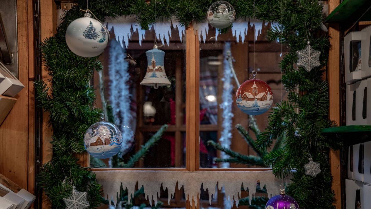 Hanging Christmas ornaments on blurred background of window framed by artificial conifer branches and tinsel. Traditional Christmas ornaments in shapes of glass balls and bells. New Year backdrop