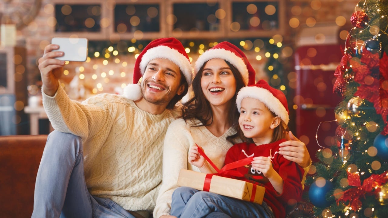 Young happy family of three in Santa hats with gifts taking selfie on phone in cozy kitchen on Christmas eve, panorama
