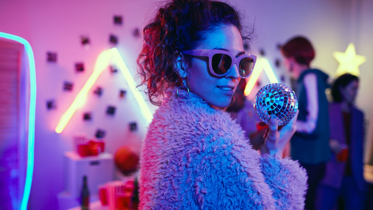 Portrait of young party girl in sunglasses and in stylish jacket looking at camera while dancing at a party