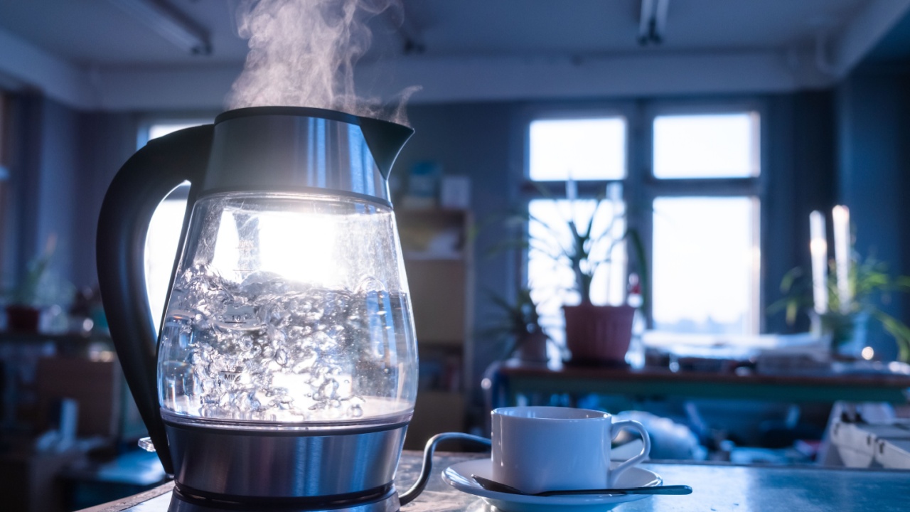 A transparent kettle of water boils against the background of the sunset shining through the window. The concept of coffee break and end of the working day.