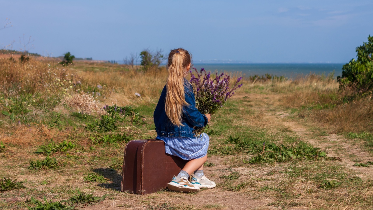 Little blonde girl in denim jacket, blue dress with vintage suitcase and flowers bouquet off-road with sea landscape. Stylish hitchhiker child with long hair on countryside trip. Kid walking outdoors.