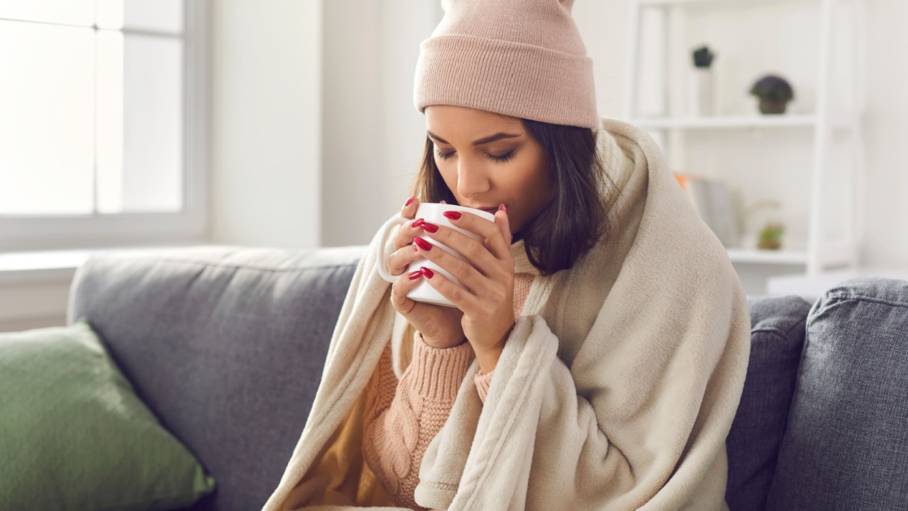 Tea to get better. Young woman wearing a knitted sweater and hat sitting at home on the sofa wrapped in a plaid drinking hot coffee or tea. Frozen woman will warm up in a cozy living room.