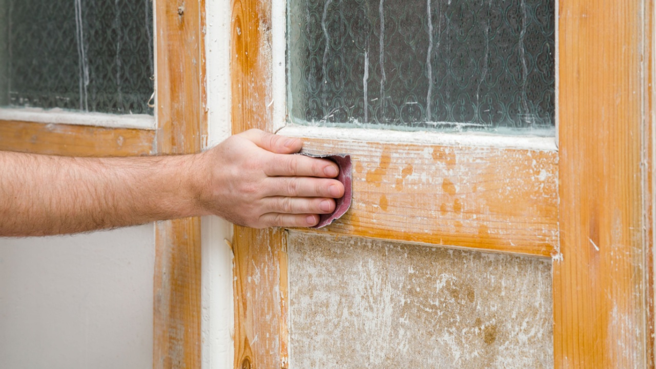 Young adult man hand using sandpaper and sanding wooden door from old white color. Closeup. Renovation process.