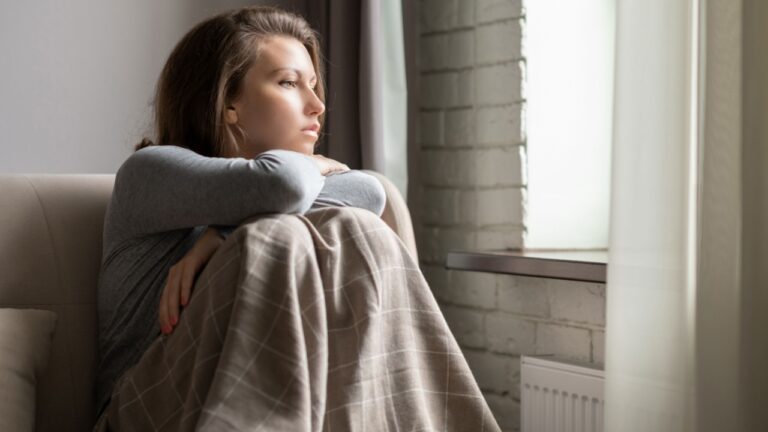Portrait of lonely sad сaucasian young woman wrapped in a plaid sitting near window of apartments.