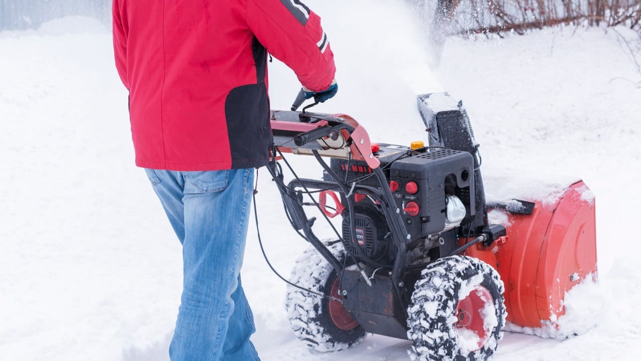 A snowblower driven by a man-driven moves along the fence and throws snow towards it. The snow-covered grassplot is cleared of snow drifts by mini-snow removal equipment. Selective focus.