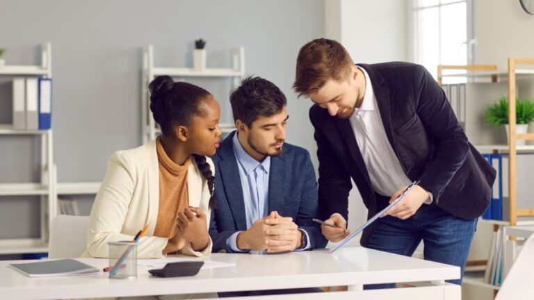 Bank employee gives documents for signature to a Caucasian man and his dark-skinned wife. Specialist in lending and financial assistance for young families indicates the place of signature.