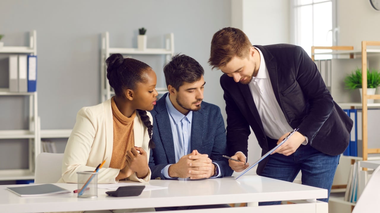 Bank employee gives documents for signature to a Caucasian man and his dark-skinned wife. Specialist in lending and financial assistance for young families indicates the place of signature.