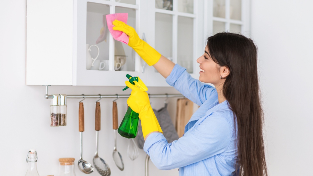 Closeup portrait of smiling woman wearing protective rubber gloves cleaning cupboard surface in modern kitchen interior, wiping dust on glass holding spray bottle and using microfiber cloth
