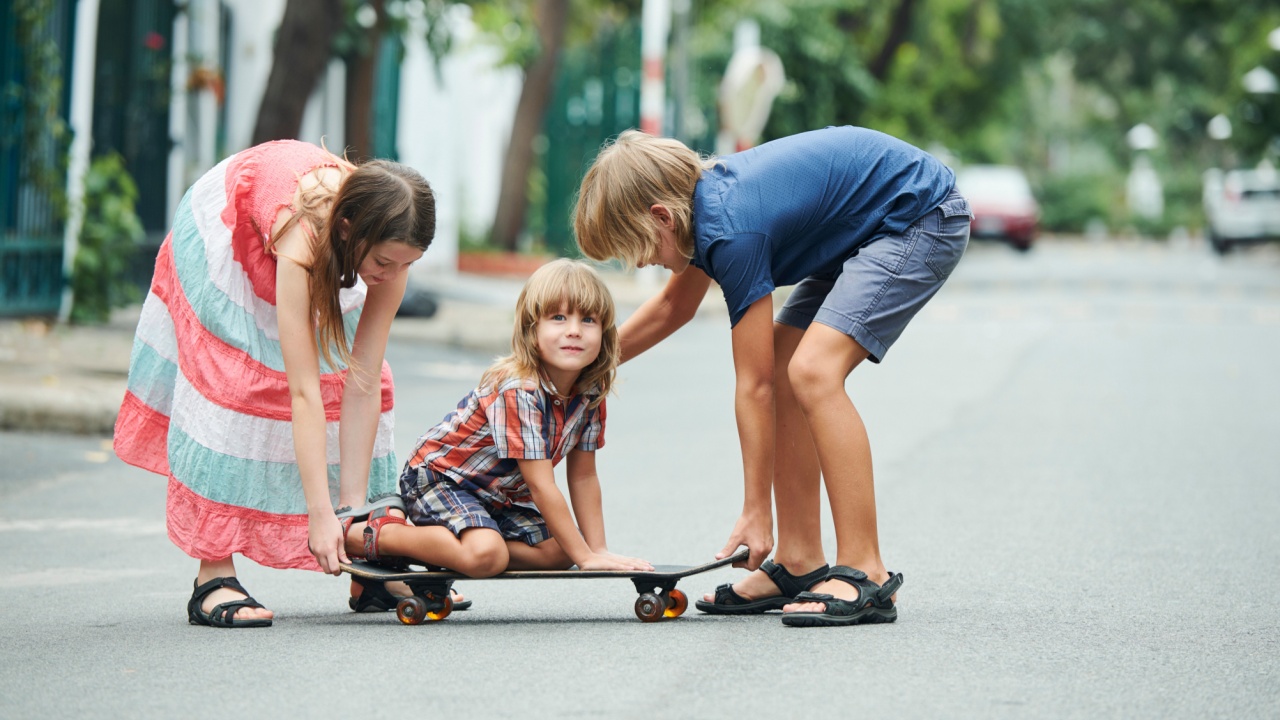 Preteen kids playing with younger brother sitting on skateboard on summer day