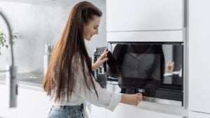 Smiling beautiful woman housewife preparing food in electric microwave oven, adjusting temperature on it. Happy female using modern kitchen built-in appliance