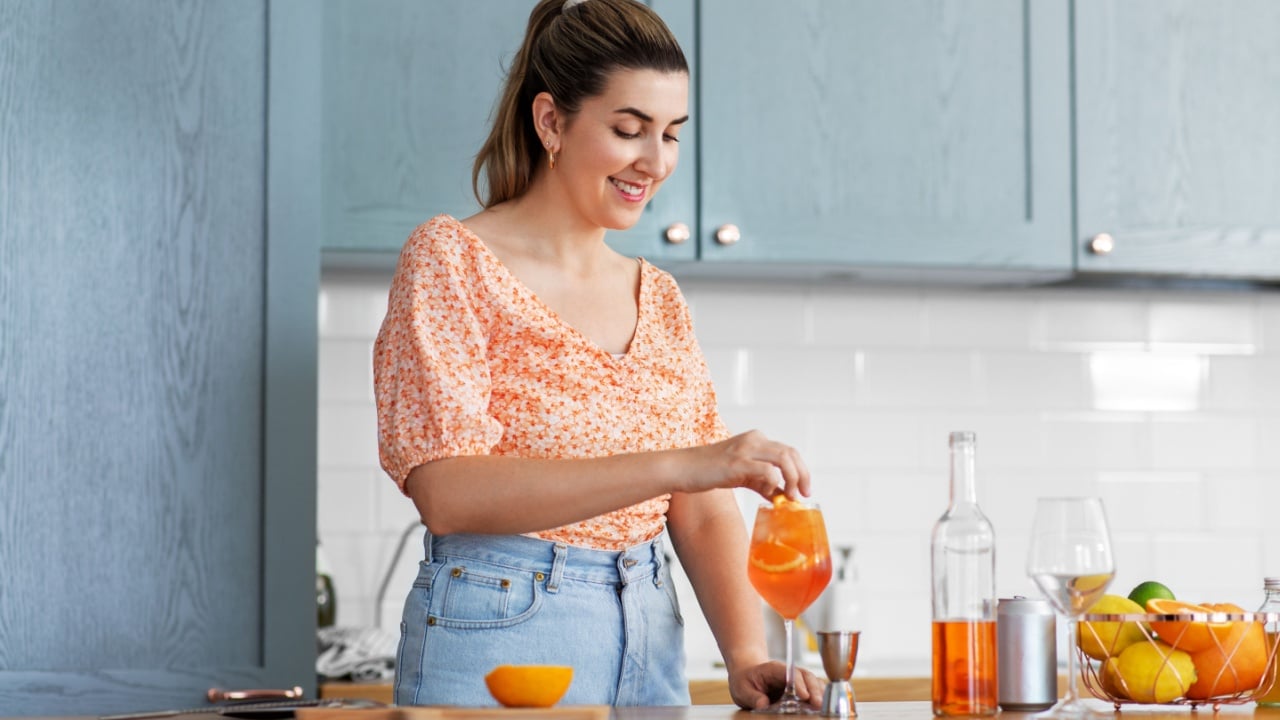 culinary, drinks and people concept - happy smiling young woman making orange cocktail at home kitchen