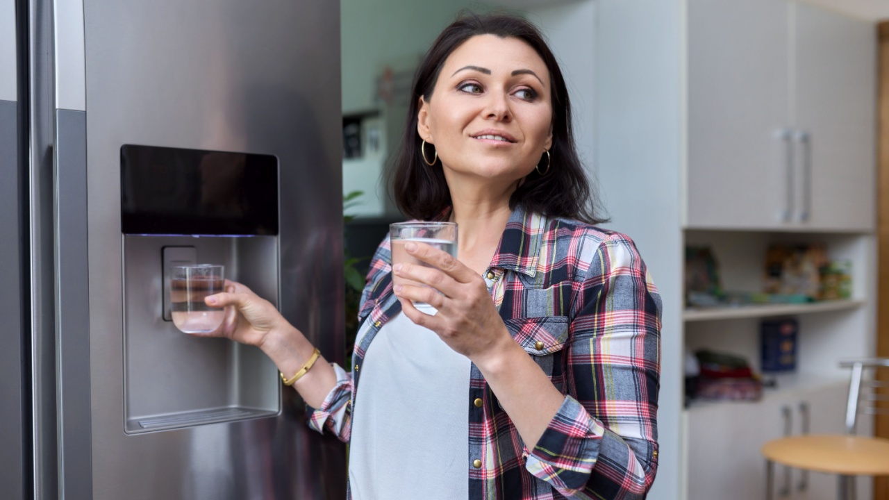 Water dispenser, woman taking cold water into glass from home refrigerator