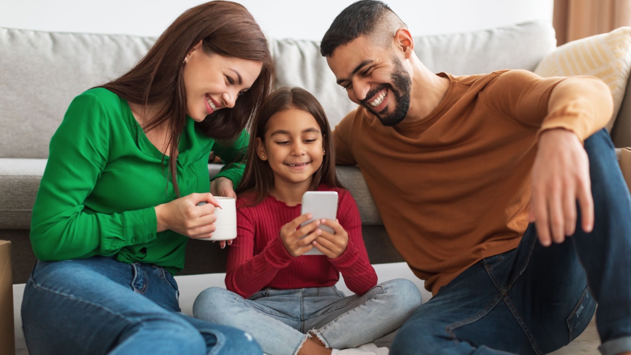 Portrait of happy young woman, man and girl holding and using smartphone sitting on the floor in living room. People spending time together, watching video, choosing gifts, daughter sharing gadget