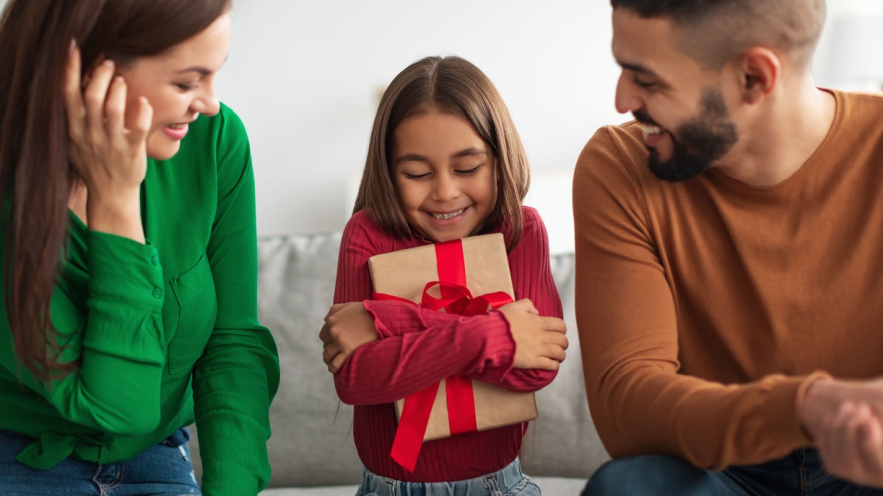 Grateful little girl sitting on couch between parents, holding and hugging wrapped gift box, man and woman greeting daughter with birthday, happy family celebrating holiday together at home