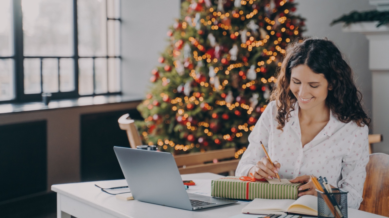 Young pleased european woman office worker writing christmas letter, greeting card with holiday wishes while sitting at her workplace with laptop and working remotely from home during winter holidays