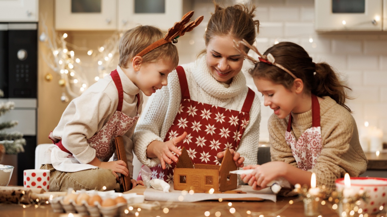 Happy family mother and two kids in xmas aprons decorating Christmas honey gingerbread house together, mom with children enjoying cooking in cozy kitchen at home during winter holidays