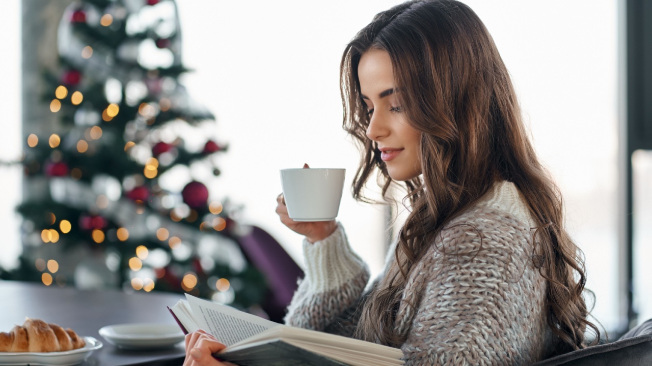Side view of young cute brunette woman in cozy sweater holding cup coffee or tea and reading interesting book on Christmas tree background. Concept of Christmas atmosphere at home.