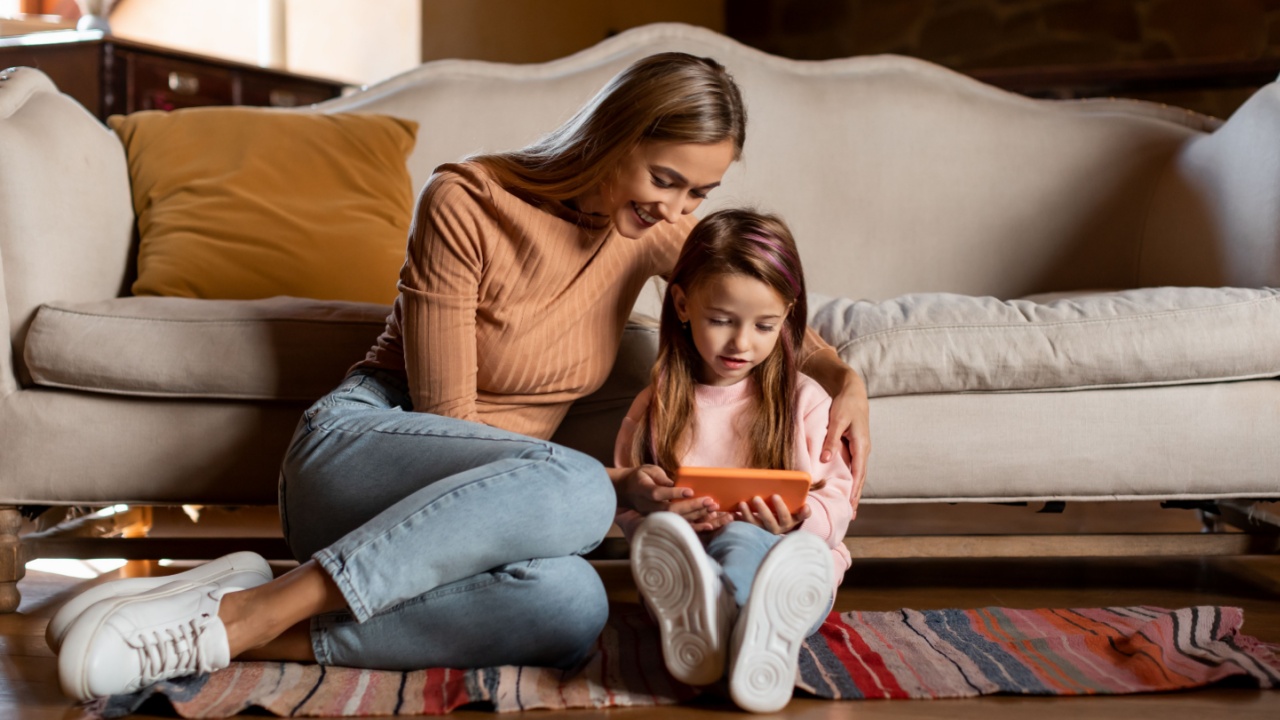 Kids And Technologies. Cheerful Little Girl Using Smartphone While Relaxing With Mom At Home, Happy Mother And Daughter Browsing New Application, Playing Gamr On Mobile Phone And Smiling, Copy Space