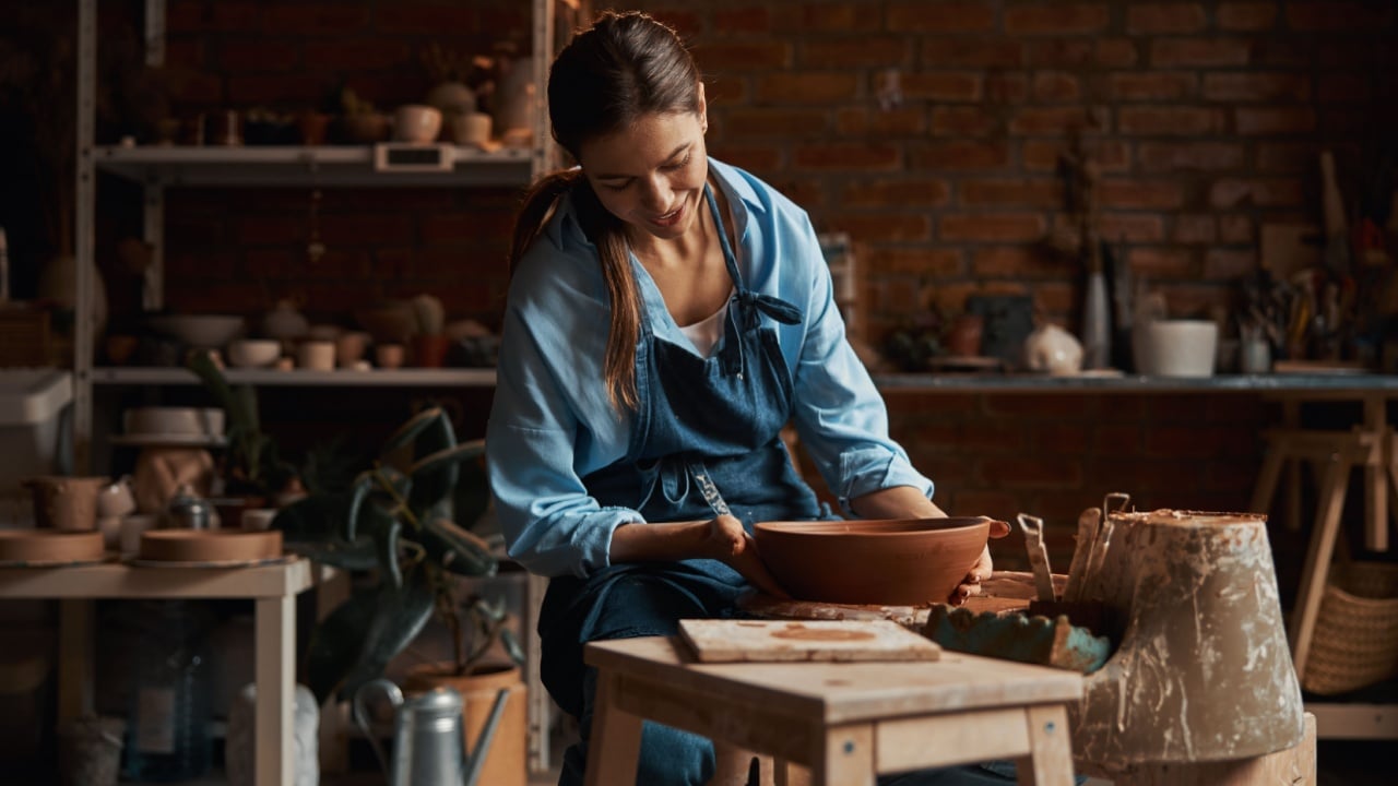 Friendly attractive female artisan wearing apron modeling ceramic tableware in pottery workshop
