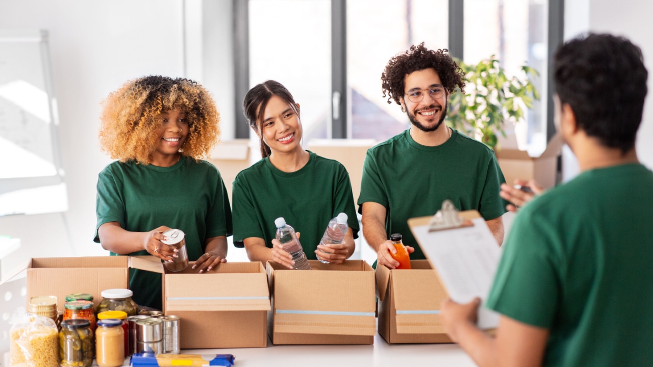 charity, donation and volunteering concept - international group of happy smiling volunteers packing food in boxes according to list on clipboard at distribution or refugee assistance center