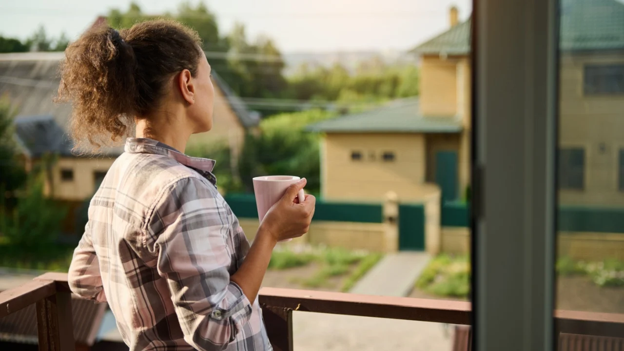 Rear view of a pretty woman in plaid casual clothes drinking coffee, admiring the neighboring residential buildings and rural scenery from the balcony of her country house on a beautiful warm day