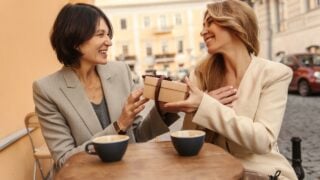 Happy caucasian adult lady on right receives holiday gift box from her friend on left. Women have nice time in street cafe with cup of coffee. People sincere emotions lifestyle concept.