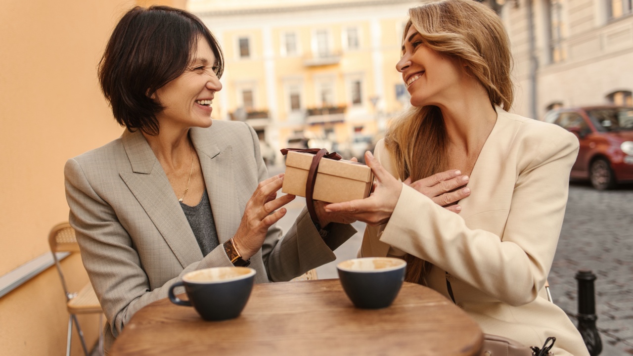 Happy caucasian adult lady on right receives holiday gift box from her friend on left. Women have nice time in street cafe with cup of coffee. People sincere emotions lifestyle concept.