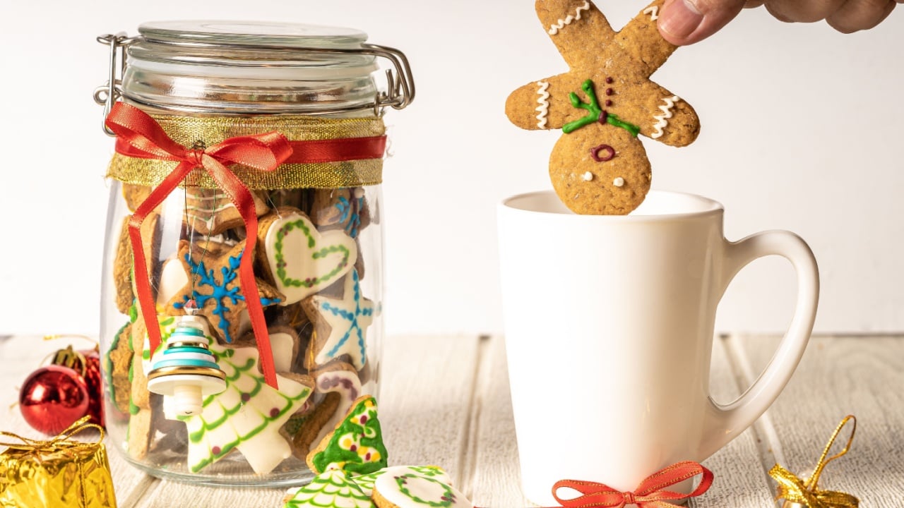 hand putting gingerbread man cookie into a mug with a christmas cookies jar on the side with white background