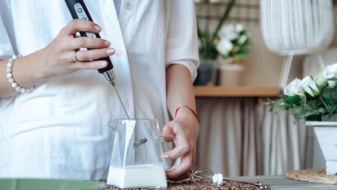 Mixing milk in glass pot by frother closeup. Female making foamy milk with handheld mixer. Housewife woman in home clothes using mixer whips cooking food in kitchen. Cook concept. Copy text space