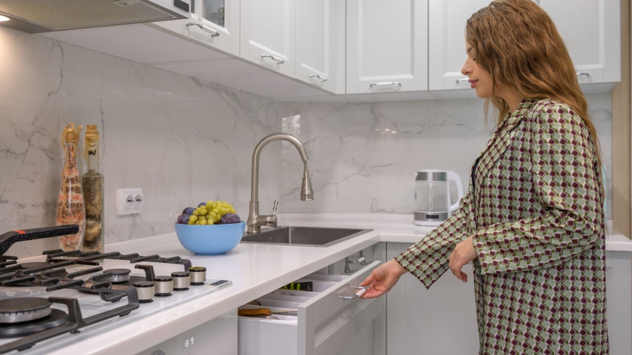 Cute young housewife pulls out the cabinet drawer at light grey kitchen