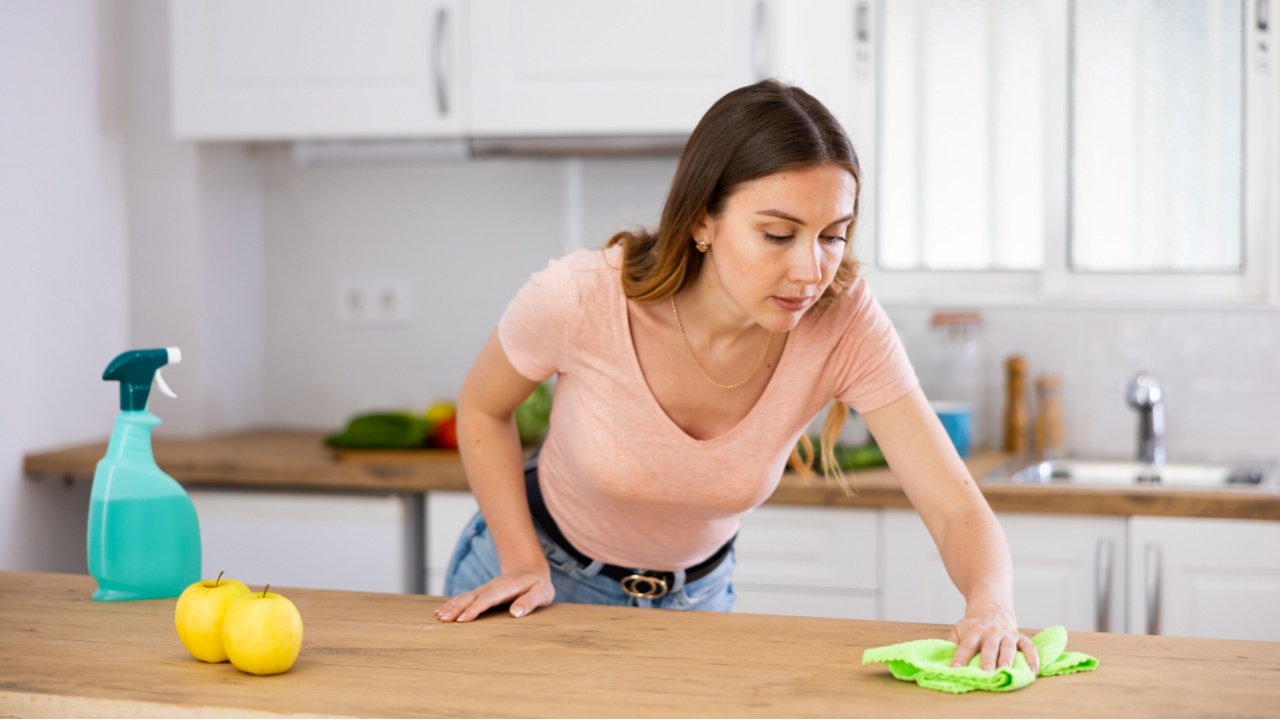 Focused woman wearing casual clothes cleaning countertop in kitchen with a rag and detergent