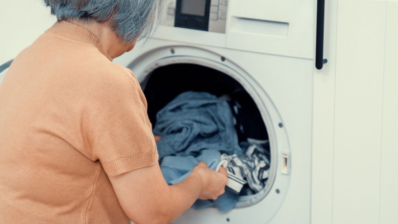 Contented senior housewife doing laundry in the laundry room with clothes inside the washing machine. Domestic life, drying machine, household chores.