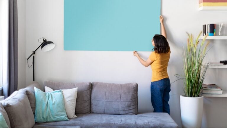 Young woman hanging large empty poster on the wall in living room at home, millennial middle eastern female decorating modern apartment with blank painting, copy space for your design, mockup