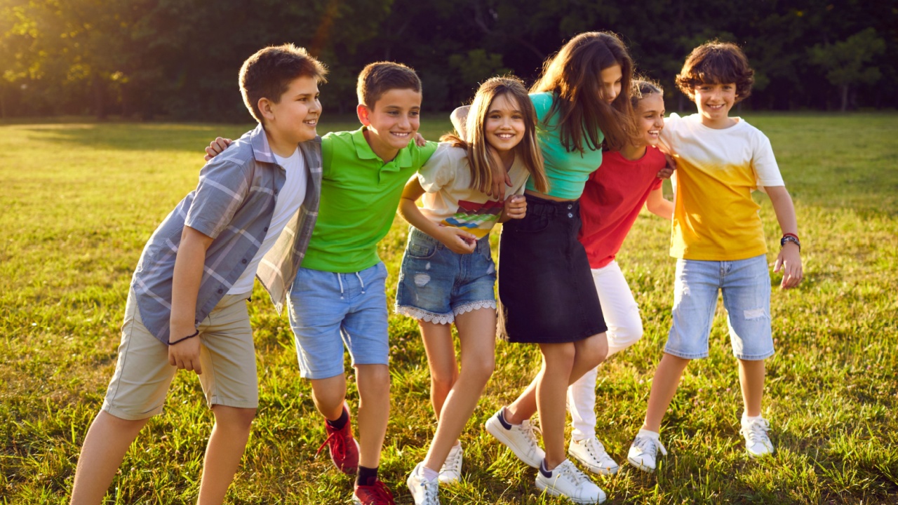 Joyful children girls and boys having good time on lawn during break between elementary school lessons smiling holding hands on shoulders of friends dressed in summer kids clothes stands in park