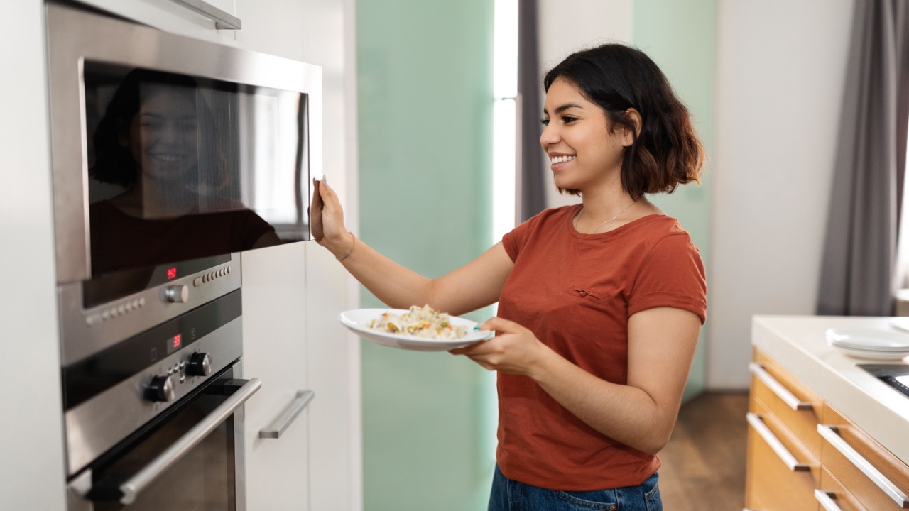 Happy Young Arab Woman Warming Food In Microwave In Kitchen, Smiling Middle Eastern Female Using Modern Appliance For Cooking At Home, Holding Plate With Meal And Opening Oven, Free Space