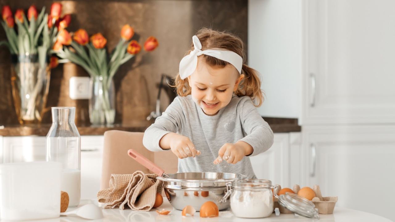 happy excited kid girl baking cake for Easter emotional child cook dessert present for mother day in white sunny kitchen Home bakery. Bottle of milk, eggs, flour are on the table