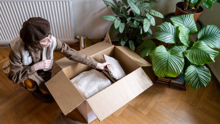 Millennial girl packing clothes for moving into new apartment, using cardboard box, donating clothing items. Young woman unpacking after move. Clothing donation concept