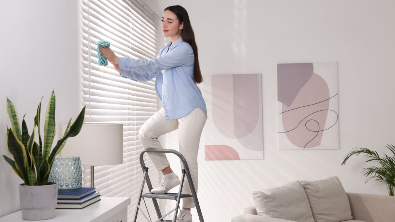 Woman on metal ladder wiping blinds at home