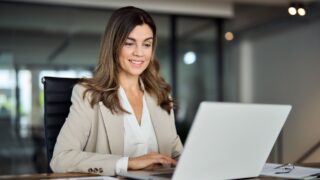 Smiling busy mature middle aged professional business woman manager executive wearing suit looking at laptop computer technology in office working on digital project sitting at desk.