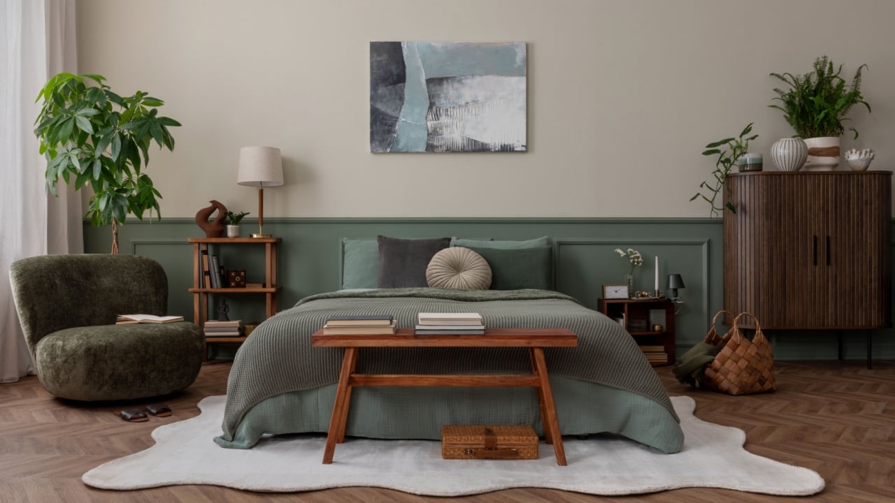 Creative composition of bedroom interior with mock up poster frame, green bedding, wooden side table, stylish bench, beige rug, plants, basket, lamp and personal accessories. Home decor. Template.