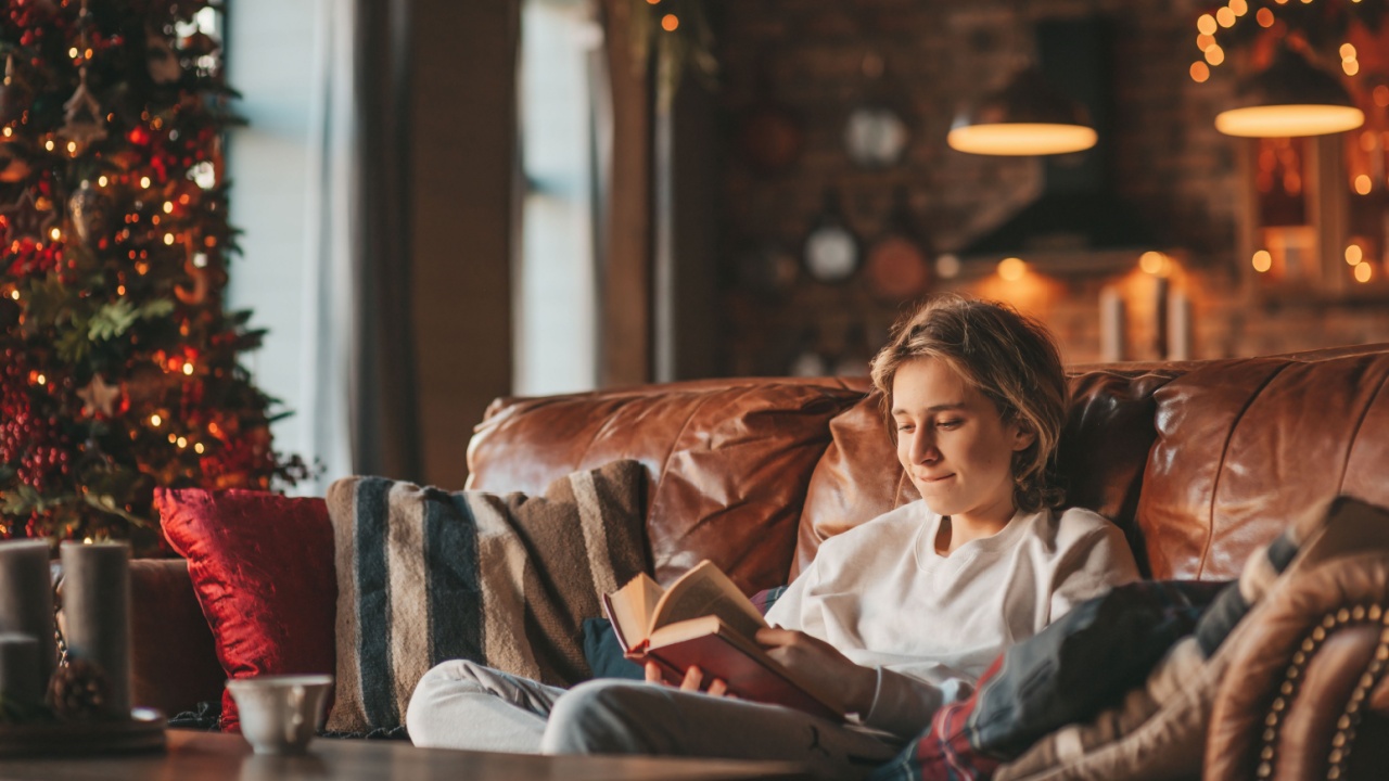 Young emotional cheerful teen boy with long hair laughter and joyful at cozy home. Stylish zoomer gen Z in good mood celebrates new year holidays with xmas tree bokeh lights garlands eve 25 december