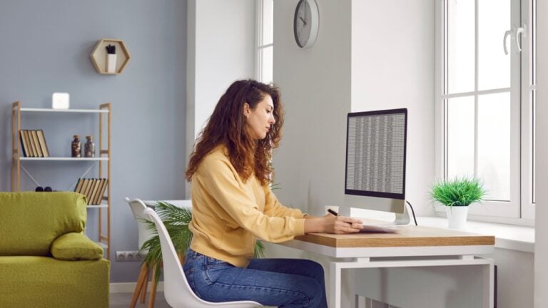 Portrait of a serious young business woman accountant sitting at the desk with pc computer with tables and charts at a modern office or at home and writing something in her notepad.
