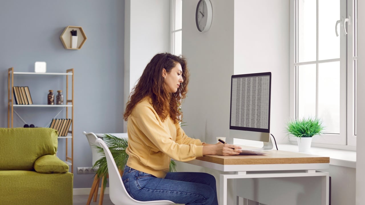 Portrait of a serious young business woman accountant sitting at the desk with pc computer with tables and charts at a modern office or at home and writing something in her notepad.
