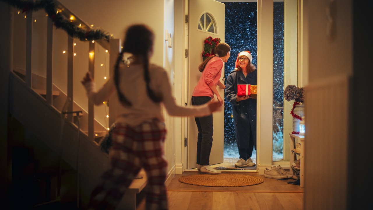 Young Mother Meeting Grandparent by the Door in a Decorated Home with Garlands, Small Boy and Girl Running to Meet Their Grandmother with Christmas Gifts and Presents