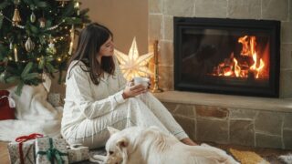 Beautiful woman in stylish pajamas with warm tea relaxing with cute dog at cozy fireplace, enjoying christmas morning in festive decorated living room with gifts. Merry Christmas! Winter holidays