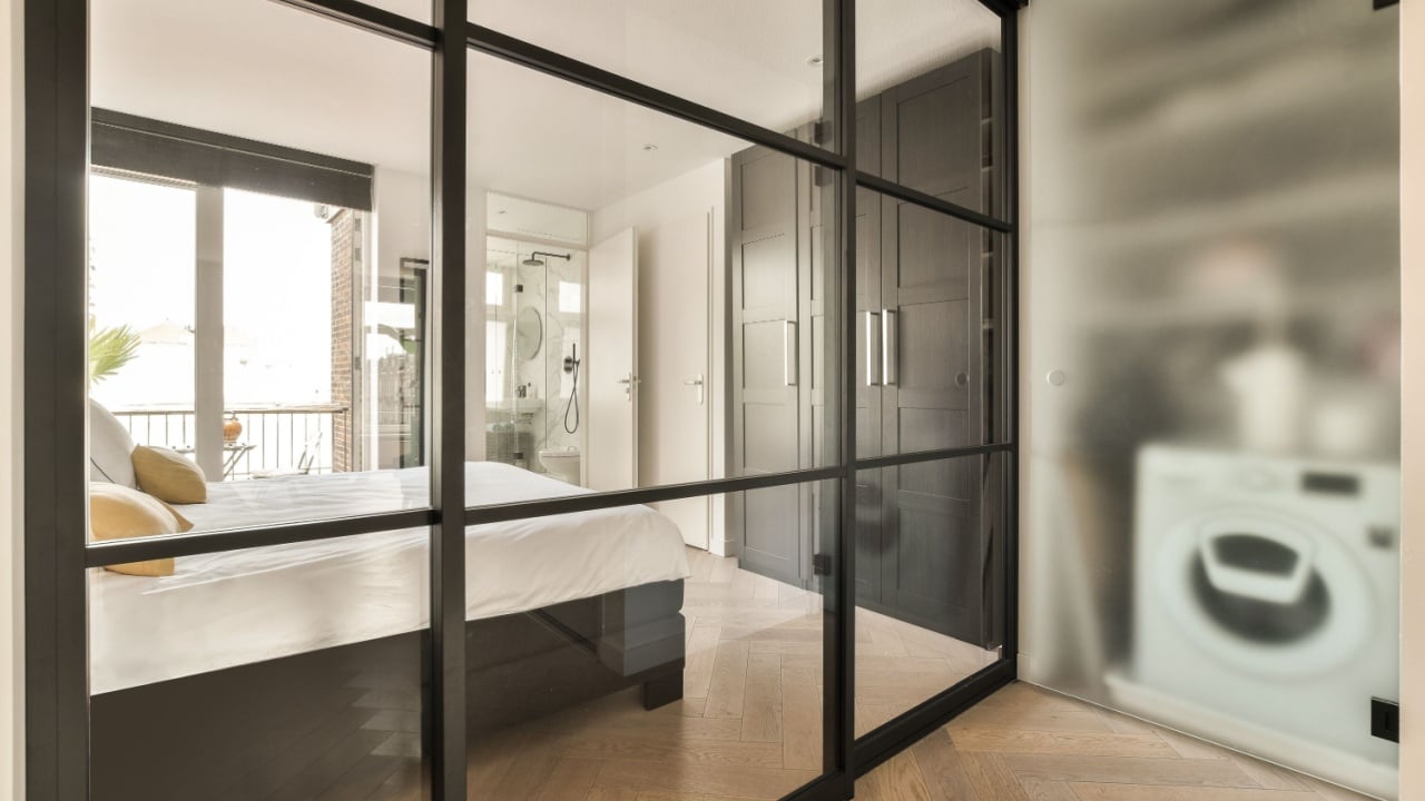 a bedroom with a large mirror on the wall, and a white bed in the room is reflected by the glass door