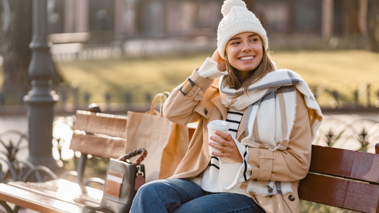 stylish woman walking in winter street wearing beige coat, knitted hat, scarf, smiling happy cold season fashion trend, shopping bag, drinking coffee