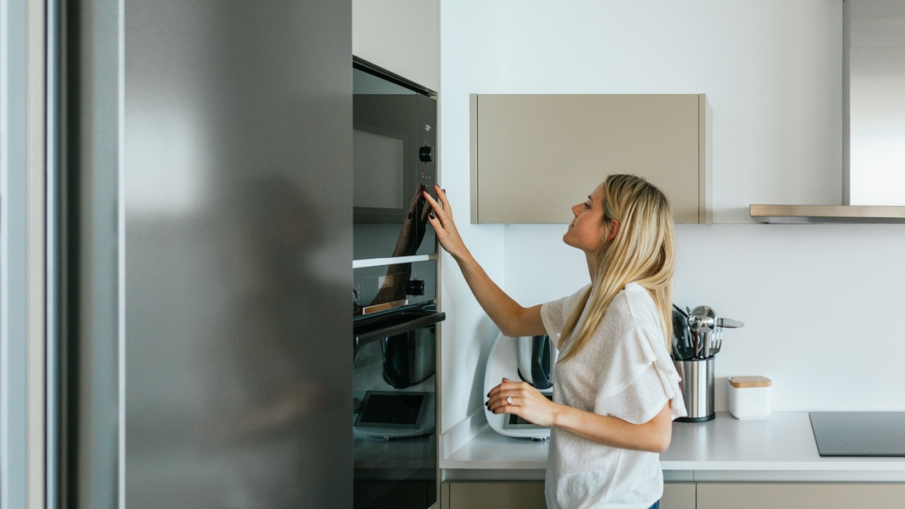 Side view of female in casual outfit turning on modern black microwave oven while standing in kitchen with modern kitchenware